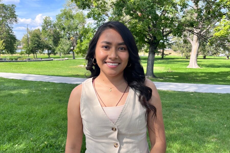 A young woman with dark hair in a beige top and white jeans smiling in a park with trees behind her.