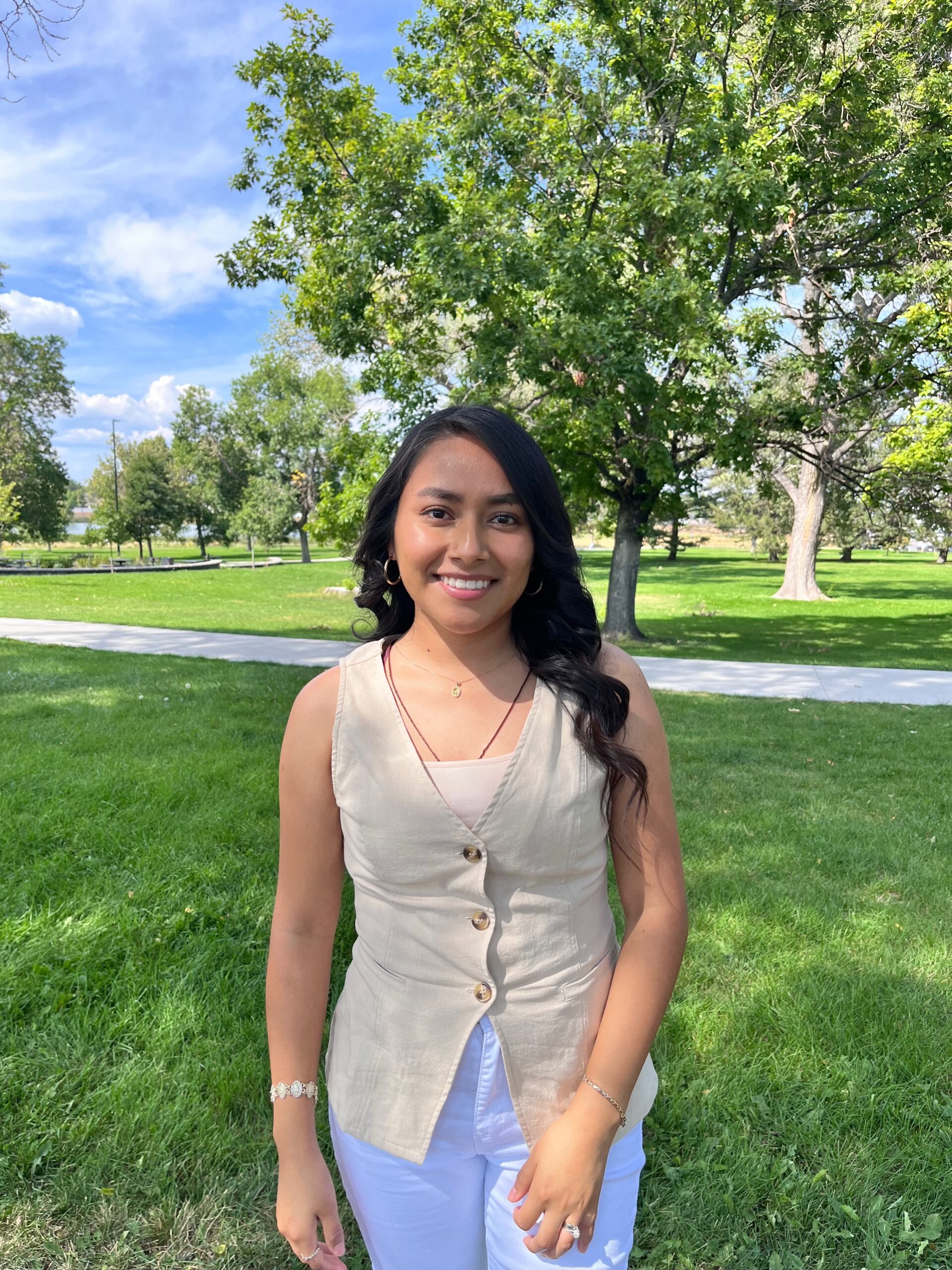A young woman with dark hair in a beige top and white jeans smiling in a park with trees behind her.