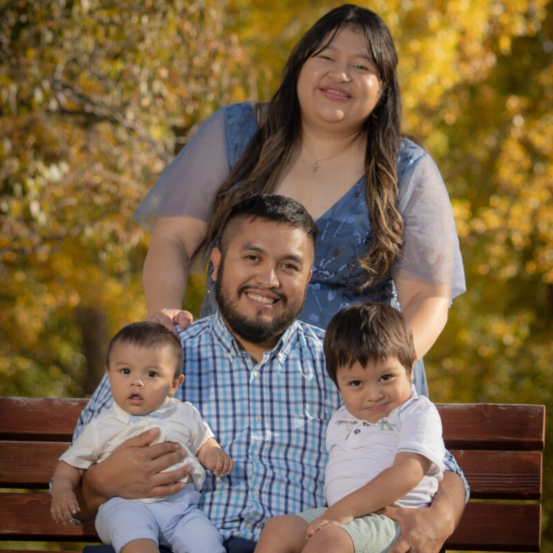 Two little boys in white being held by their father in a blue checkered shirt, with their mom standing behind them in a blue dress.