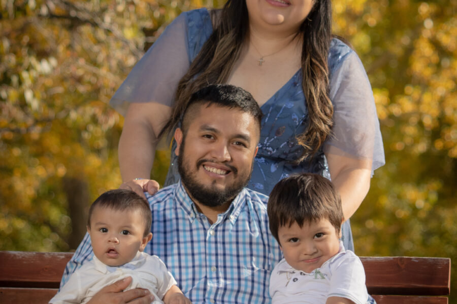Two little boys in white being held by their father in a blue checkered shirt, with their mom standing behind them in a blue dress.