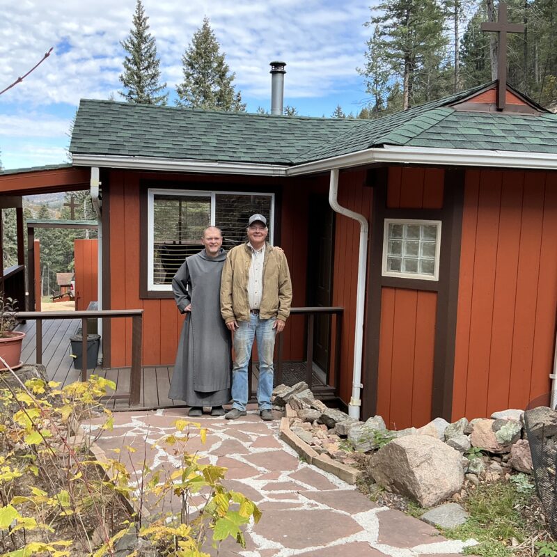 A man in jeans and a brown jacket and a religious brother in a gray habit stand in front of a red cabin in the Rocky Mountains.