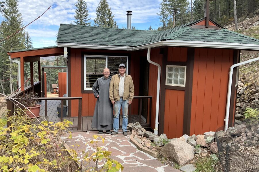 A man in jeans and a brown jacket and a religious brother in a gray habit stand in front of a red cabin in the Rocky Mountains.