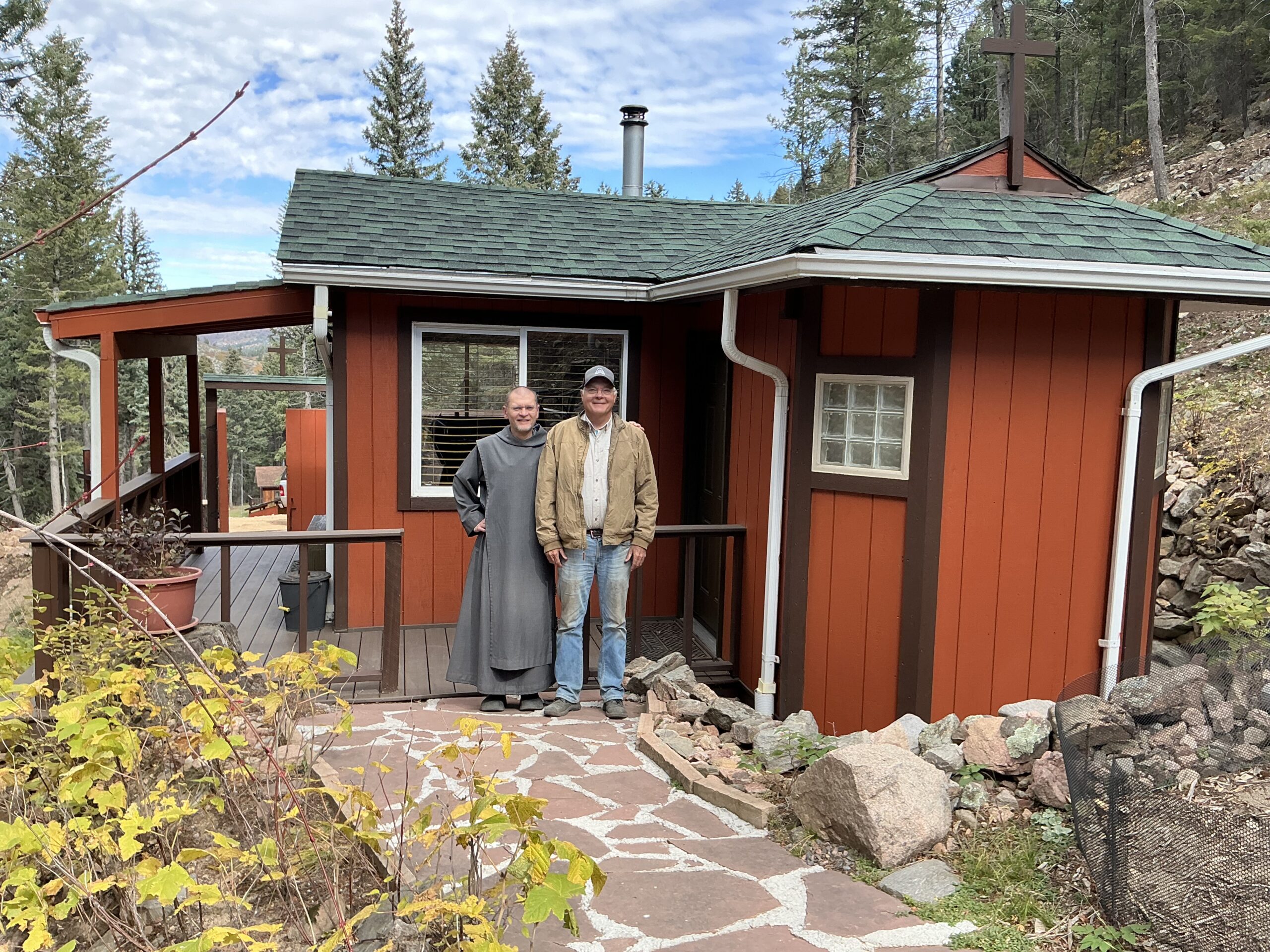 A man in jeans and a brown jacket and a religious brother in a gray habit stand in front of a red cabin in the Rocky Mountains.