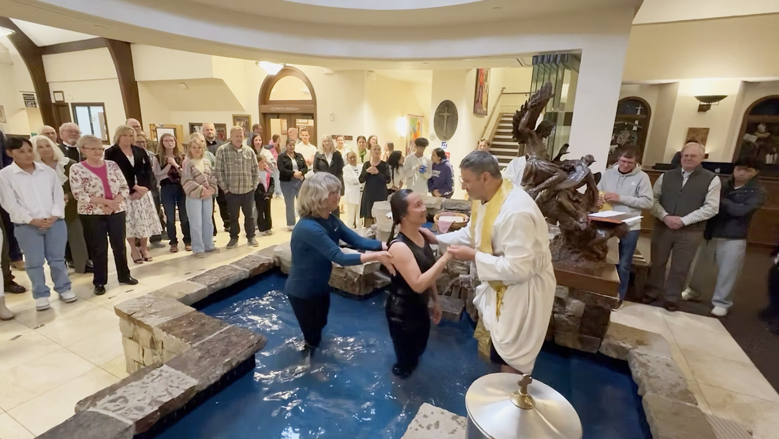 A woman dressed in black being baptized in a stone baptismal font by a Catholic priest.